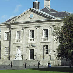 Photograph of a wide two storey grey building with a blue clock near the top. The building has steps leading up to it and a white statue in the front