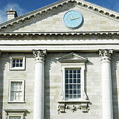 Photograph of a grey building with a large window in the middle of two columns. At the centre top of the building is a large blue clock.