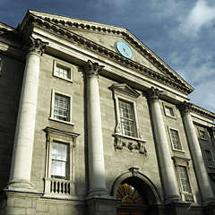Photograph taken from ground view of a tall old grey building with 7 windows and a blue clock at the top centre of the building and an archway at the bottom
