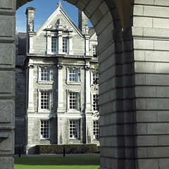 Photograph of a tall grey building with a pointed roof and two visible chimneys. The building is photographed through an archway.