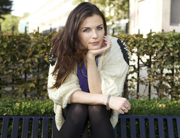 Image of Aisling Bea sitting in front of a hedge on a sunny day.