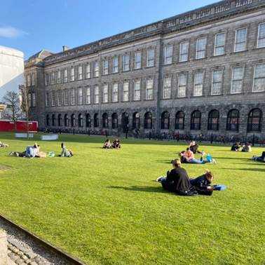 A group of people sitting on the grass in front of a large building  Description automatically generated with medium confidence